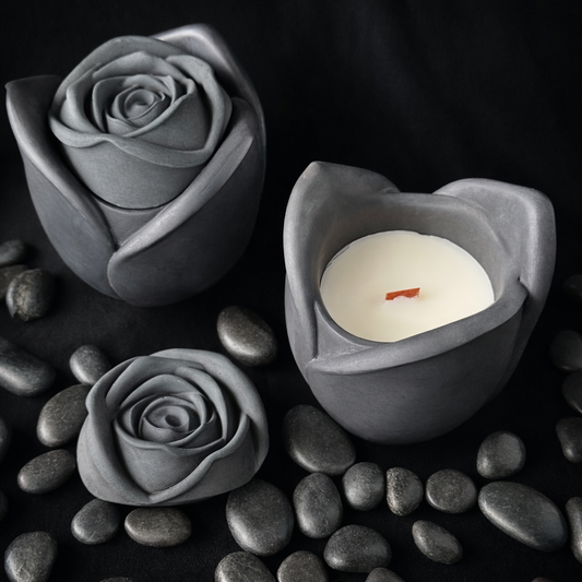 Black colored rose-shaped cement candle holders with a white soy paraffin blend wax wood wick candle on black pebbles. Goth black rose candle 