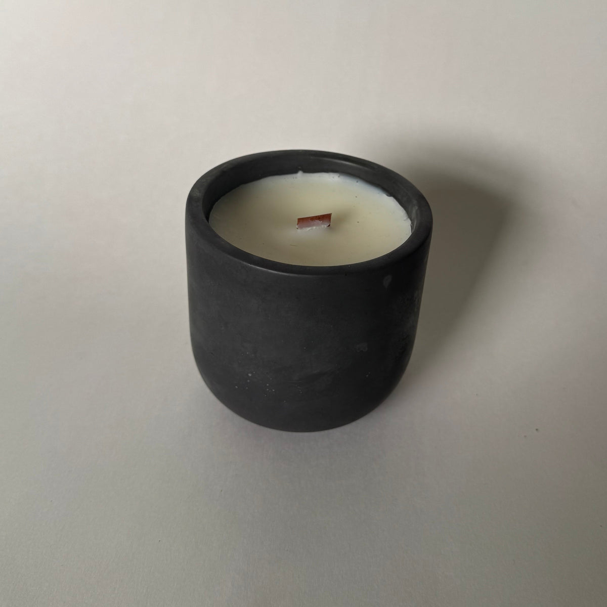 A minimalist black cylindrical candle holder with white wax and a single wooden wick, set against a plain light gray background.