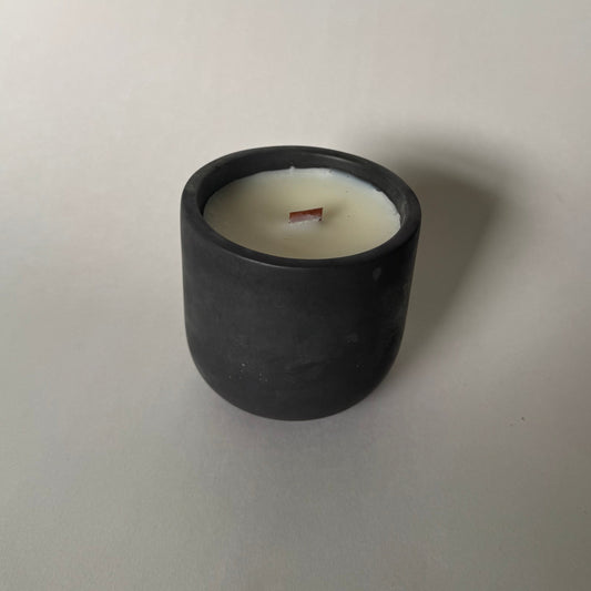 A minimalist black cylindrical candle holder with white wax and a single wooden wick, set against a plain light gray background.