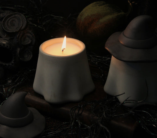 Three matte gray cement witch hats candle holders arranged around a lit white candle on a dark, spooky background with a skull and dried leaves.
