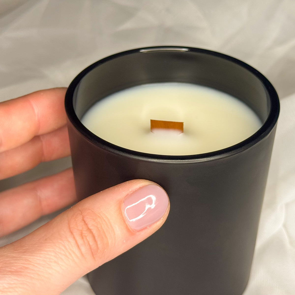Hand holding a black candle with a wooden wick against a neutral background