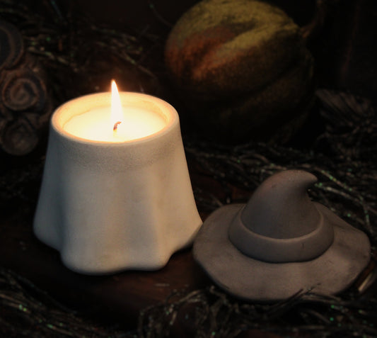 A white candle holder shaped like a ghost lit with a warm flame, displayed next to a small gray witch hat candle holder on a dark, moody background with dried leaves and a pumpkin.